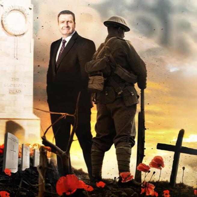 John Nichol in a suit stood next to a World War I soldier facing away from him, a cenotaph, war graves and crosses as you would see on top of a grave. Poppies appear at their feet.