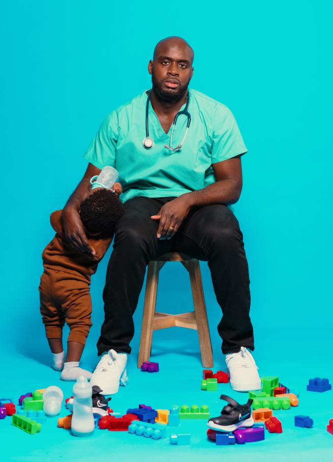 Comedian sitting on stool accompanied by small child holding a milk bottle.