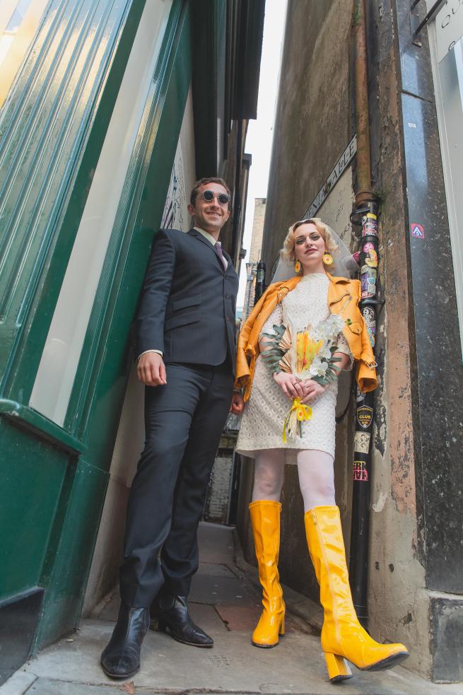 50s looking couple standing in an alleyway, one with yellow boots and jacket and short midi wedding dress, the other in navy blue suit and glasses