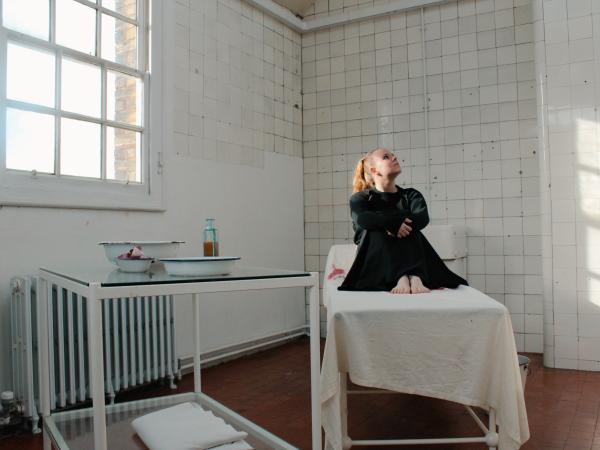 Playwright and musician Amanda Grace sitting on a bed in a tiled Victorian hospital.