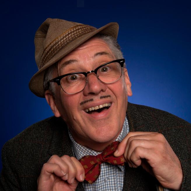 Arthur is wearing a hat, glasses, a tweed jacket and a red bowtie. He has a skinny moustache and is smiling at the camera. The background is blue.