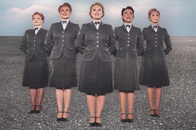 Five members of the cast of D-Day Darlings stood on tarmac in an arrow formation wearing grey blazers, bow ties and knee length skirts.