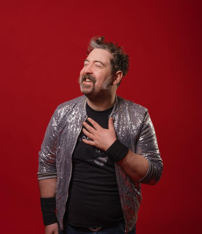 Nick Helm in a silver sequin jacket with his right hand on his chest looking up to the left.