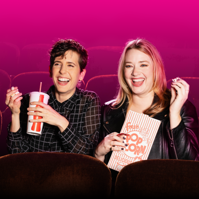 Two women sat in a cinema auditorium. laughing. One holds popcorn and the other holds a drink.