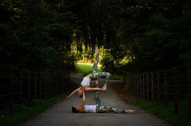 Two ladies, performing acroyoga on a park path, with trees arching overhead. They in mid pose, with one holding the other up with a foot and hands, the woman being held is gracefully arching backwards