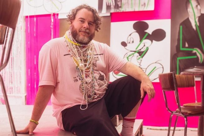Person with a beard in a pink t-shirt with layers of beaded and pearl necklaces sitting relaxed in a pink room. 