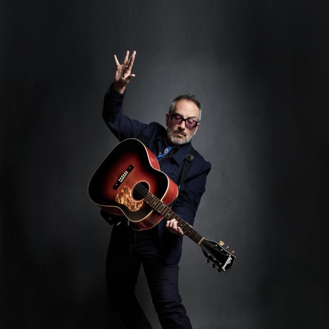 Elvis Costello with an acoustic guitar looking down on the guitar where his left hand is on the neck with his right hand up in the air.