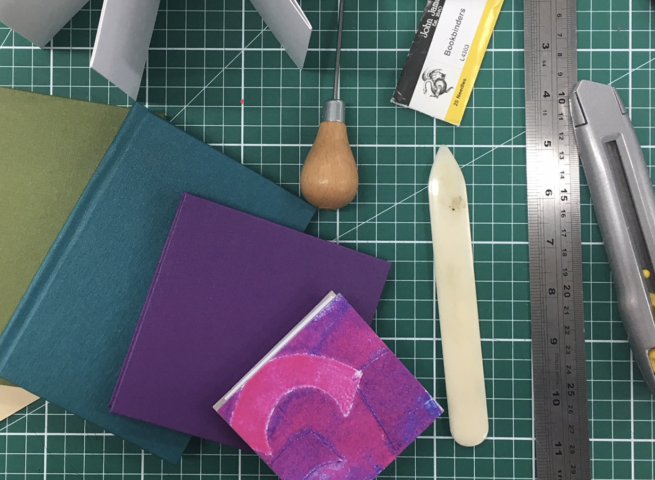 The image shows some small coloured notebooks lying on a green cutting mat, along with some bookbinding tools and equipment.