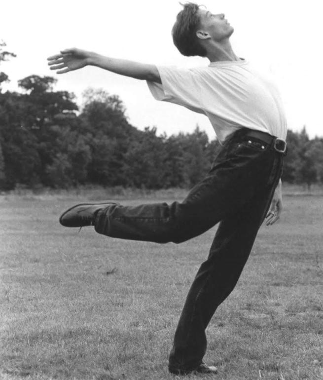 A black and white image of a young male dancer aged about 24 in a field grass under foot and trees in the background. The dancer is dressed in a white t-shirt and black jeans. His body is arched like a bow and his front right leg is bent behind him with his foot at knee height trailing behind and his front right arm reaching backwards just above shoulder height as he looks upwards at the sky.