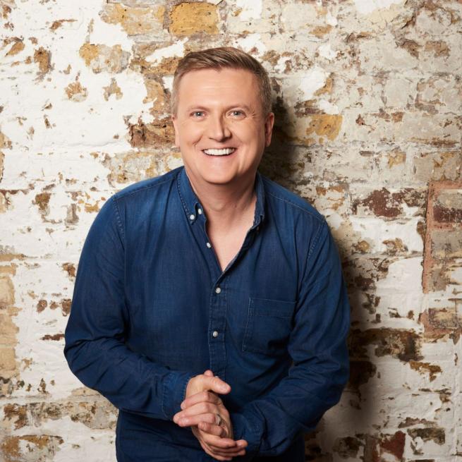 Aled Jones stood against an exposed brick wall smiling with his hands clasped together below his chest.