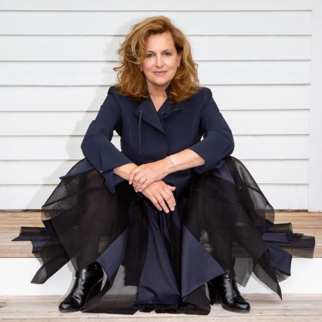 Barbara Dickson in a navy suit jacket and a tulle skirt sat against a white slatted wall.