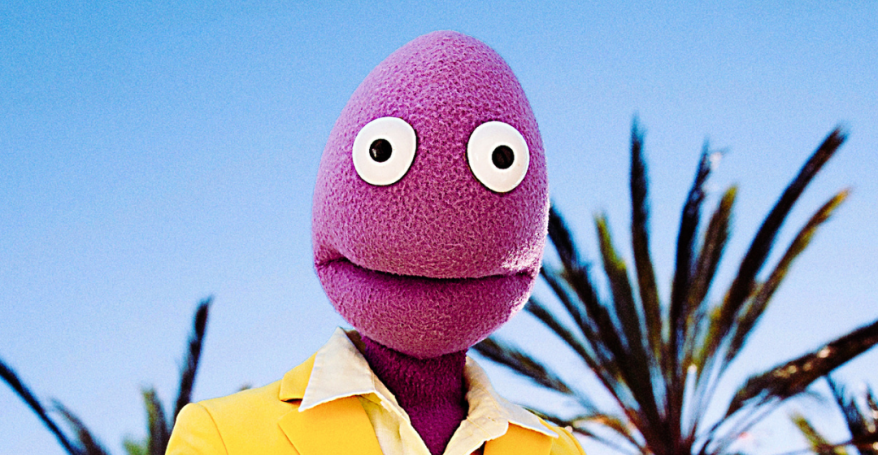 Randy Feltface pictured from the chest up wearing a bright yellow suit and his purple hands to his chest. His purple felt face and round eyes are staring right out to the camera in the centre of the shot, Blue sky and palm trees appear in the background.