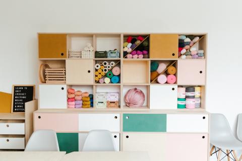Photograph featuring colourful studio shelving full of craft materials next to a big wooden studio table and grey chairs 