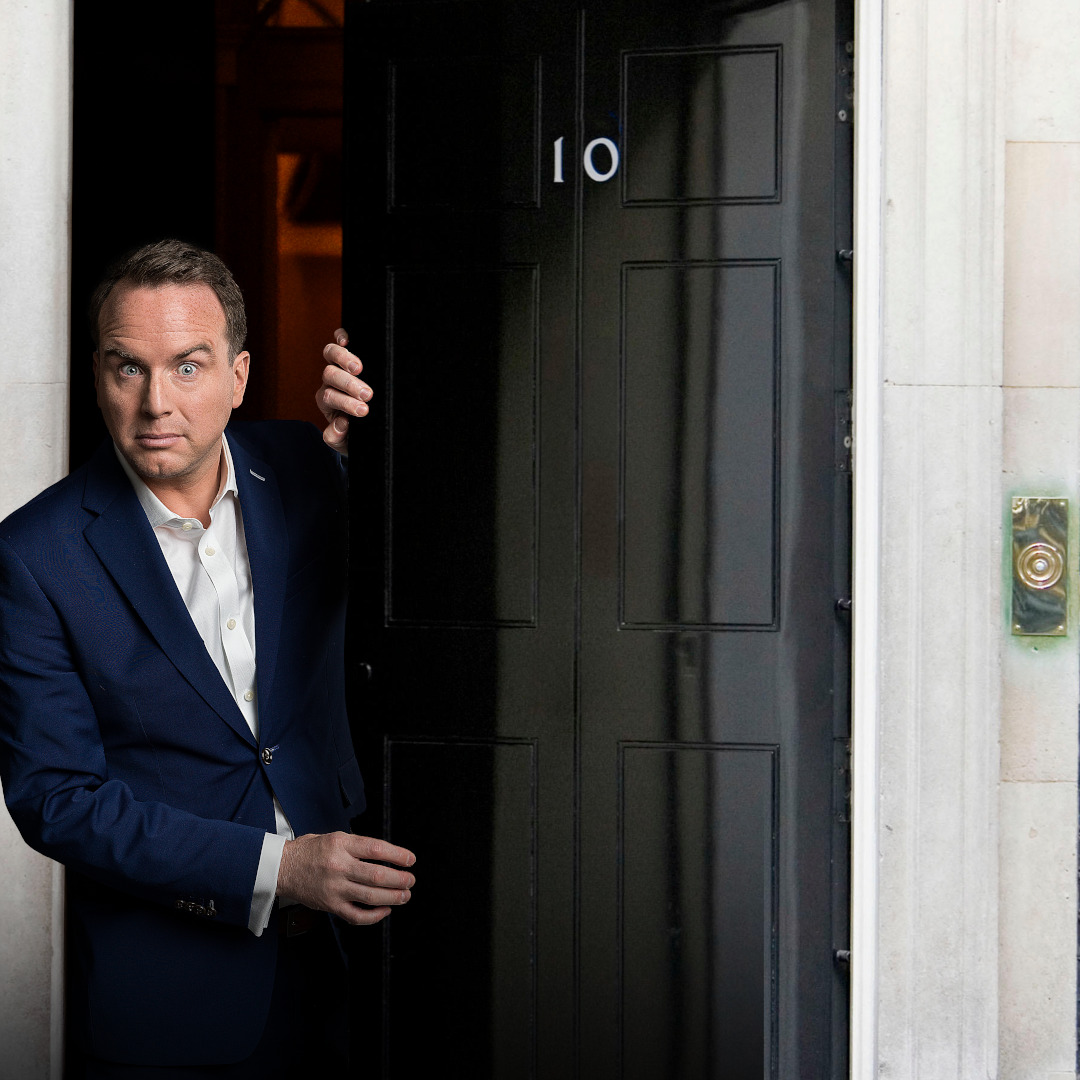 Matt Forde is appearing out of a house and opens the door. He is standing next to a black door with the number 10 on it.