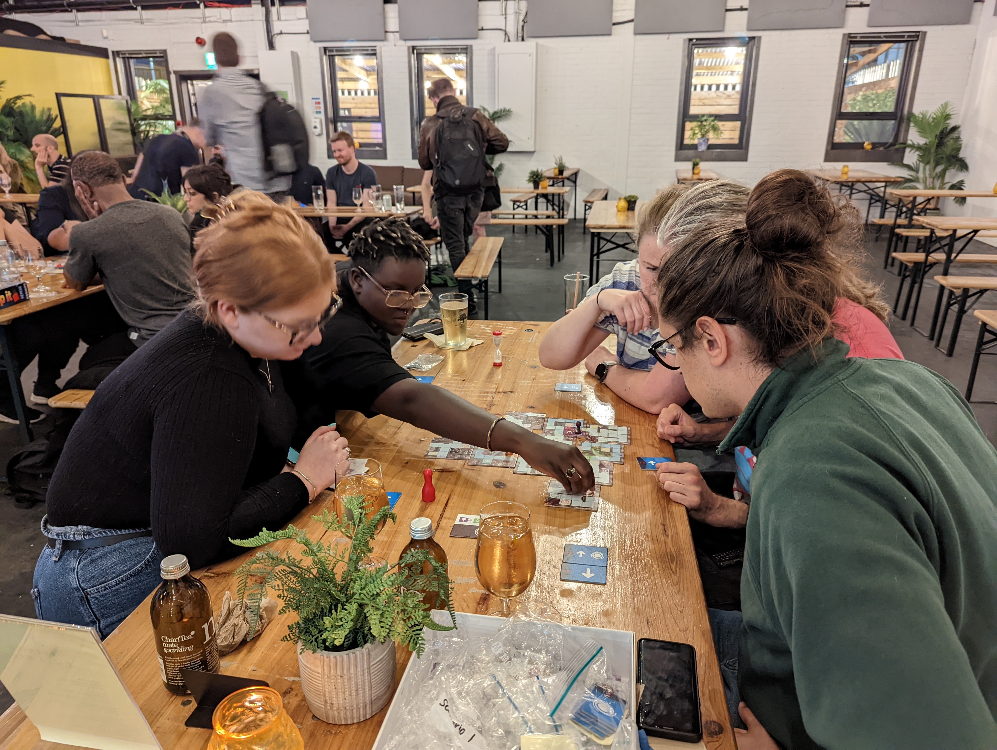 Players are seated round a large table. They are leaning in, all focussed on the board game they are playing. One player is leaning across and moving a piece. 