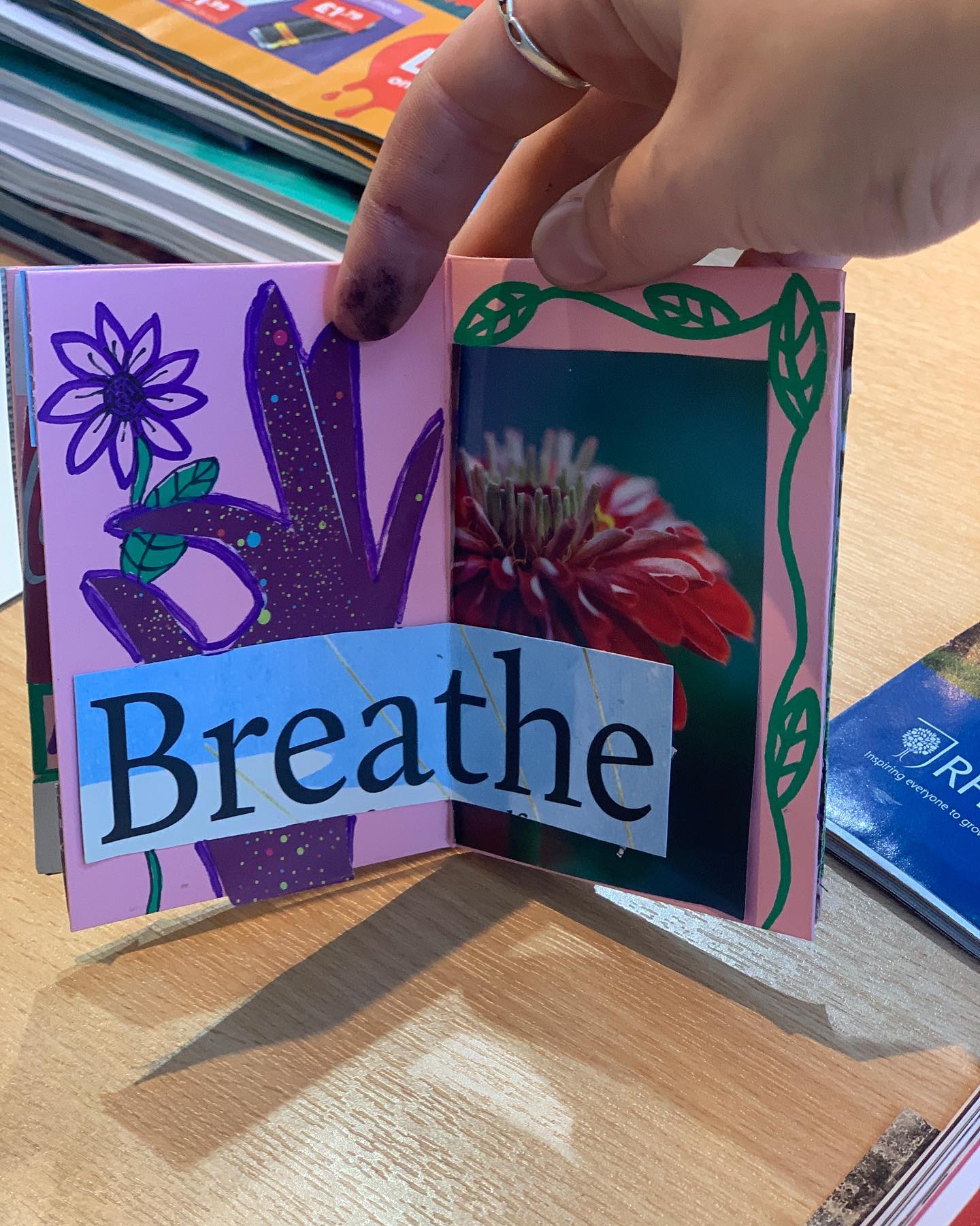 A hand holds up a pocket-sized booklet, collaged with the word 'breathe' and pictures of flowers.