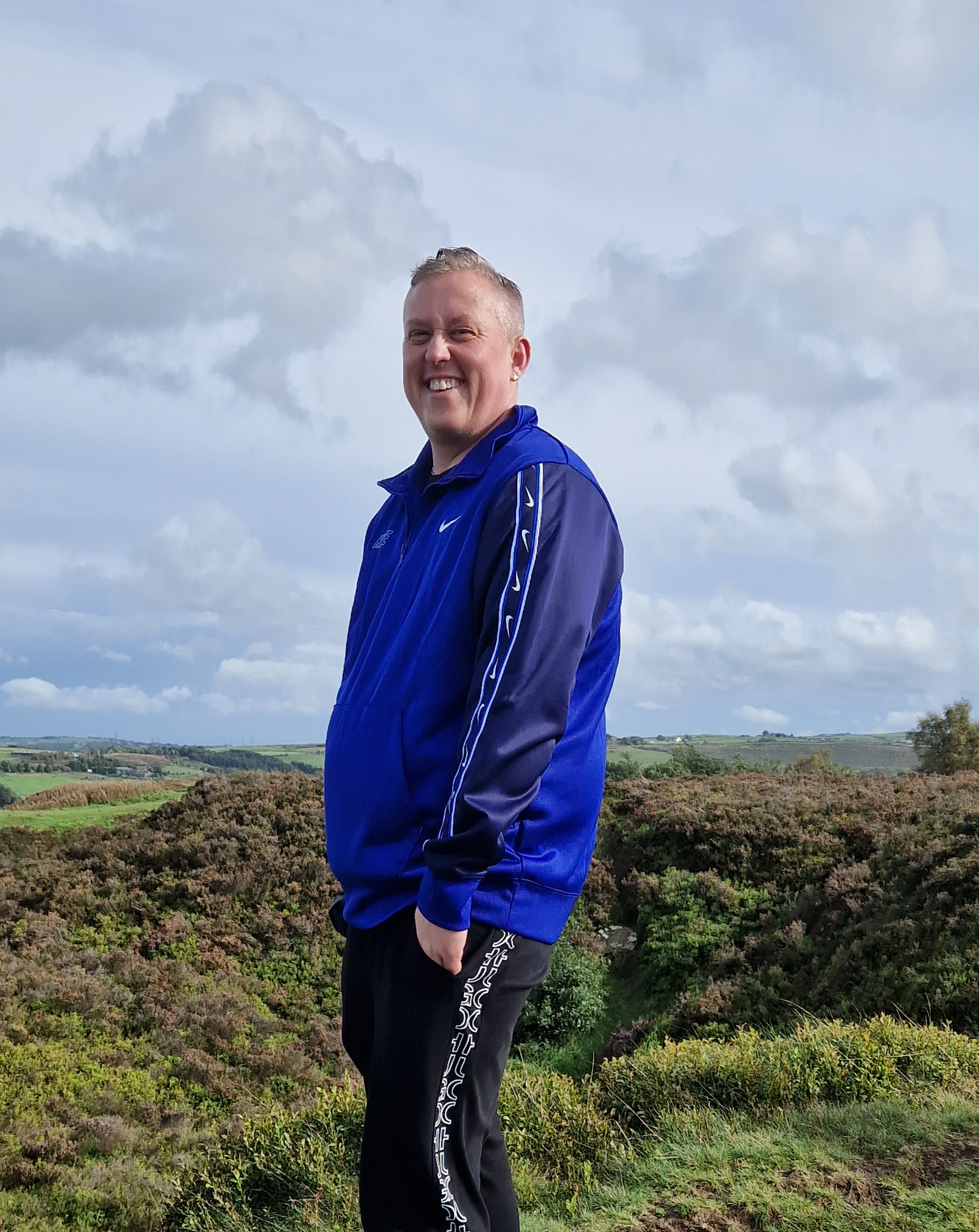 A white transgender man in a blue and black tracksuit stood on a moor.