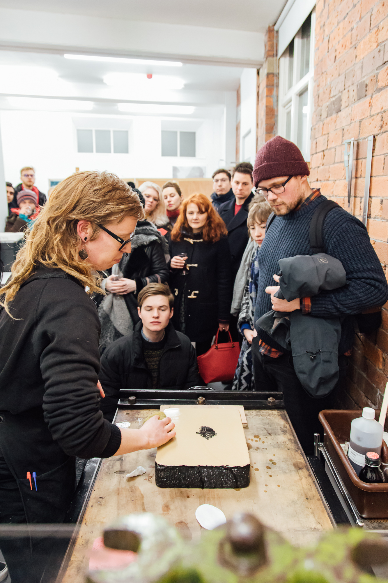 Artist demonstrating how to use a printing press which an audience surrounding 