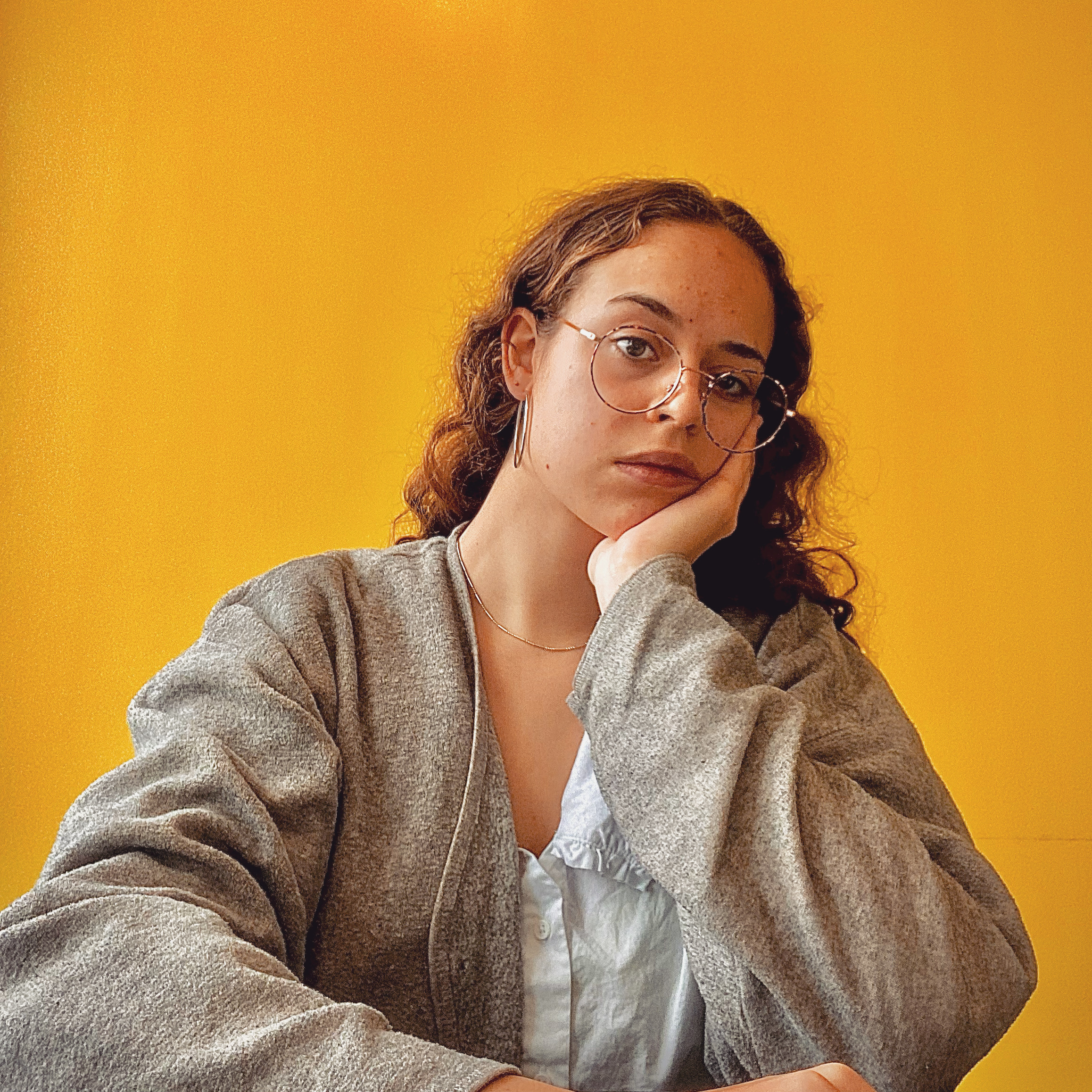 HerOrangeCoat main image: the artist with her hand on her chin against an orange background. She is wearing a grey cardigan and white shirt, and has glasses and curly hair.