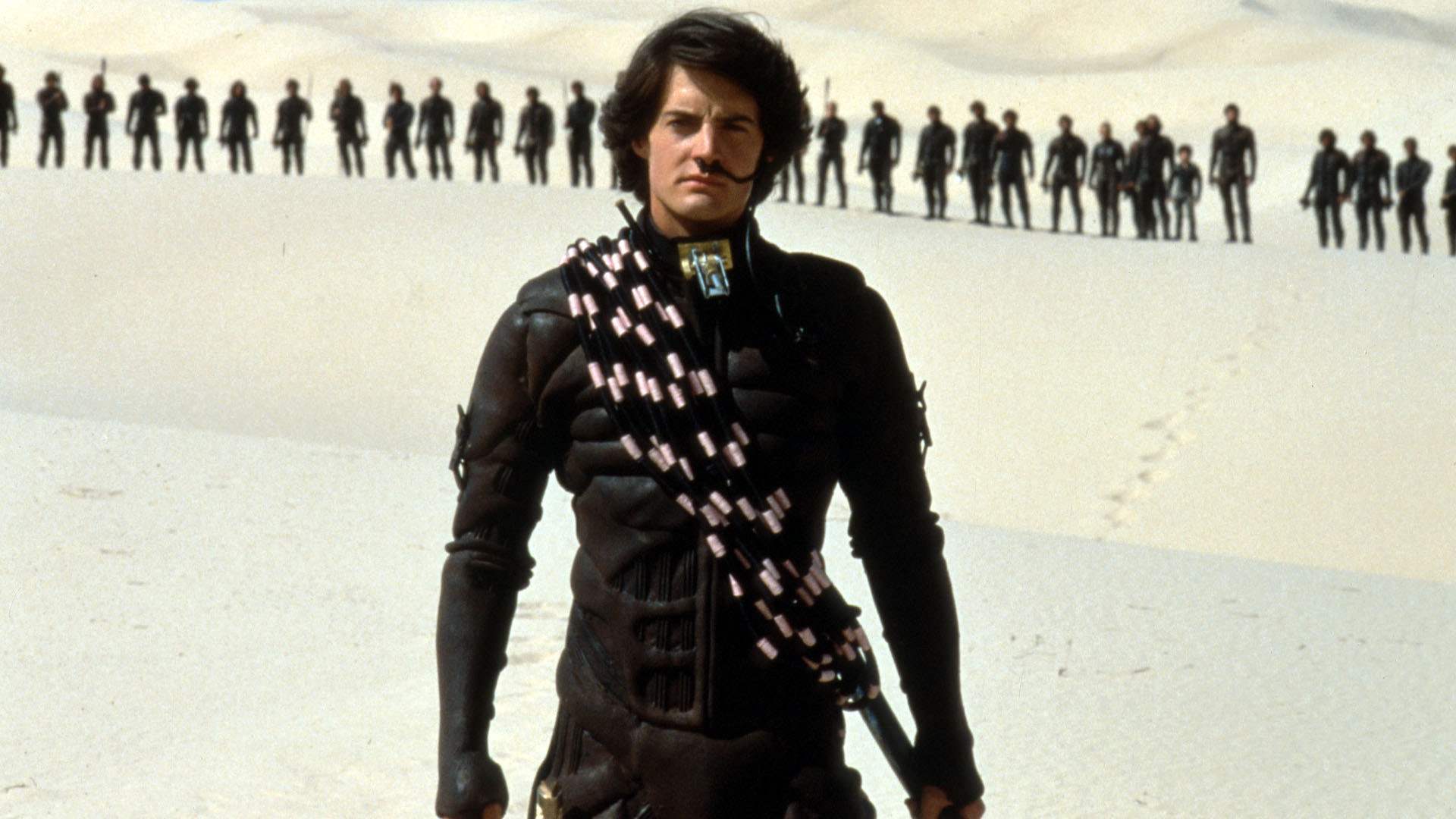 A man in an all-blac outfit and a black breathing apparatus in his nostril is in front of a line of similarly dressed people in a white desert.