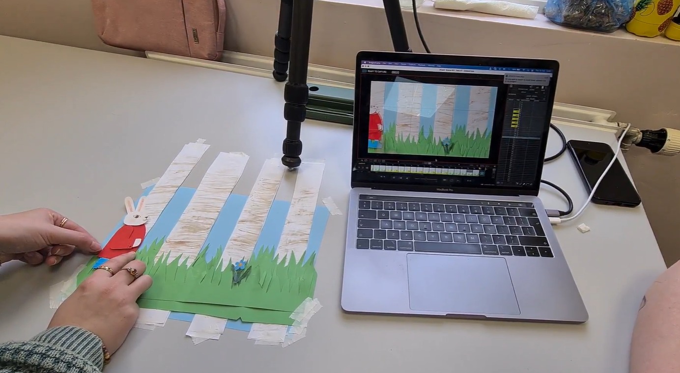 Image of a white desk with a pair of hand operating a stop motion rabbit paper puppet next to a laptop. The stop motion scene can be seen duplicated on the screen of the laptop. 