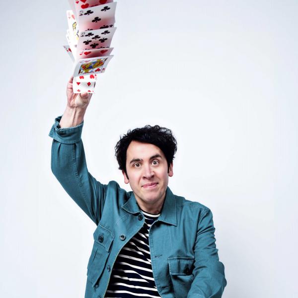 Pete Firman in a striped shirt with a jean jacket overtop. He has playing cards in his hands and is beginning to toss them into the air.