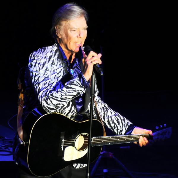 Against a black backdrop, Marty Wilde sings into a microphone holding a guitar.