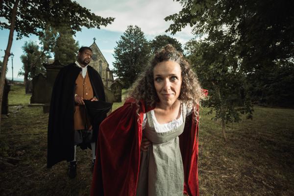 Woman wearing red cape smirks directly into camera. Man in black cape, holding tricorn hat, lurks in the background. The pair are surrounded by trees in a church yard.