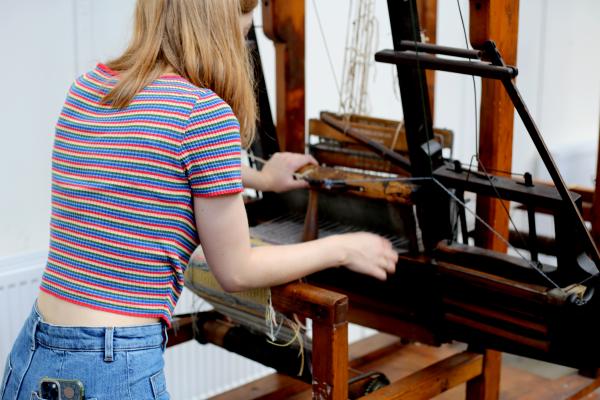 A young woman stands at a dark wooden loom with her hands at work. She is wearing a rainbow stripe top.