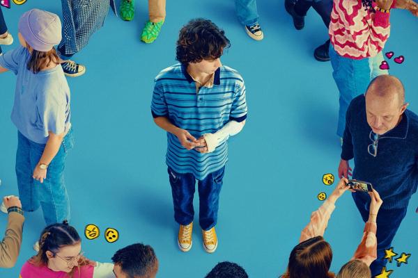 Evan Hansen in a stripy polo shirt with a cast on his arm, stood surrounded by other people on a blue floor.