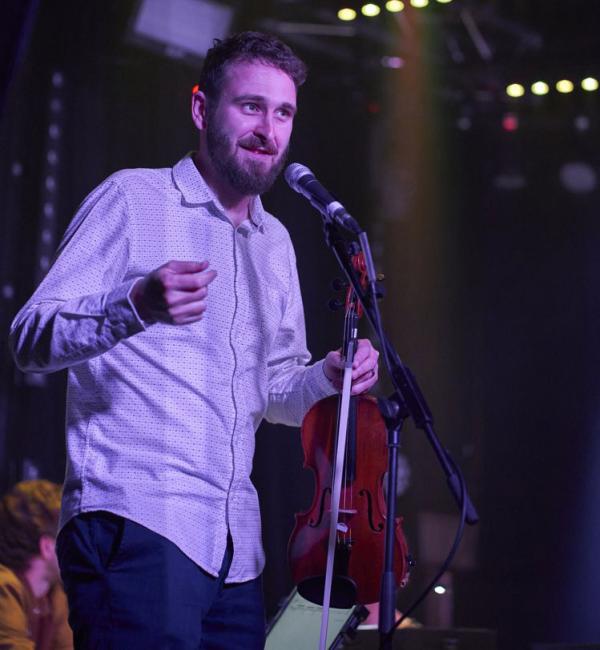 A man standing talking into a microphone holding his violin. 