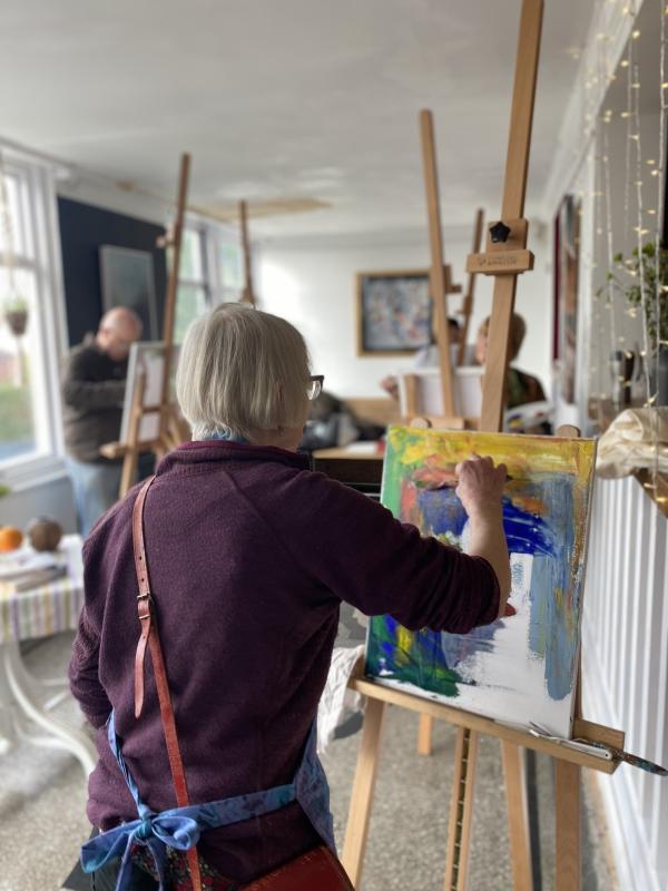 A woman stands at her easel painting a canvas using a palette knife. 