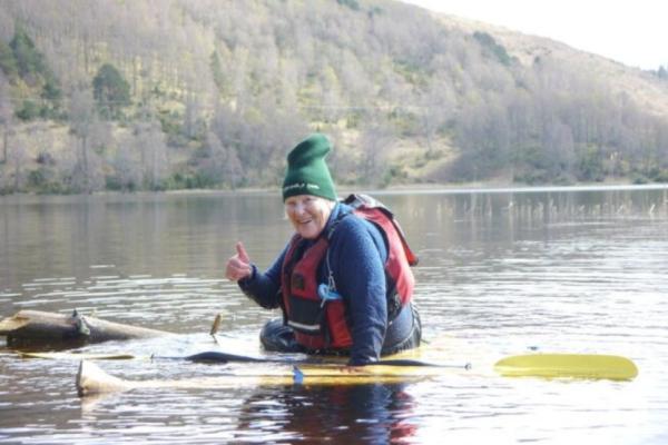 An elderly person in a forest green beanie and red life jacket on a yellow kayak on a peaceful lake, surrounded by tree filled mountain landscape. They are giving the camera a thumbs up.