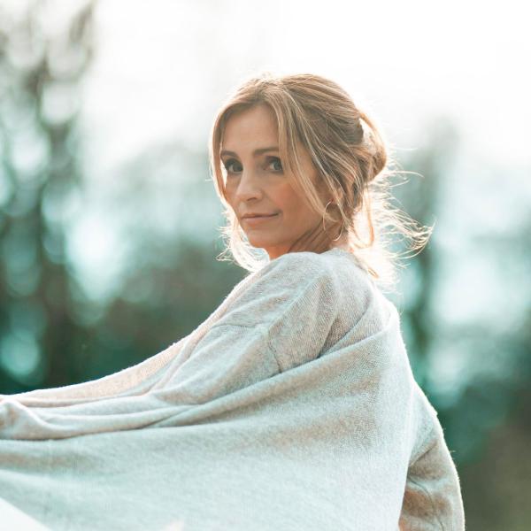 Cara Dillon from the shoulders up wearing a pale grey cardigan looking over her left shoulder back at the camera. Blurred out trees appear in the background.