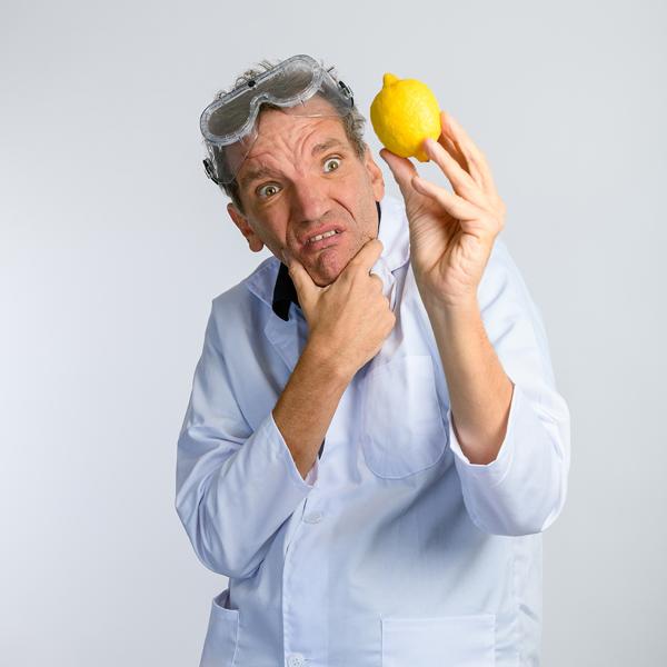 Henning Wehn in a white lab coat and safety goggles on his head holding a lemon in his left hand looking at it perplexed.