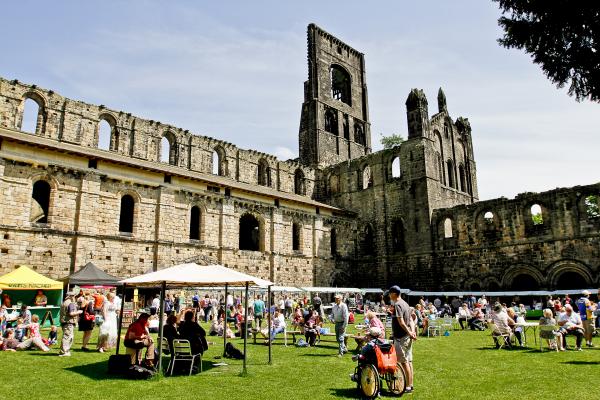 kirkstall market on a sunny day, stalls surround a grassed area with lots of people sat around drinking and eating with the abbey set as the backdrop.