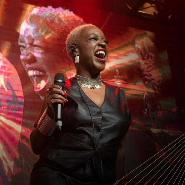 Woman looking very happy on stage wearing a black leather waistcoat and holding up a microphone in her right hand. She appears on the screen behind her.