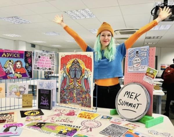 A photograph showing artist Jenna Greenwood, with a sample of some of the artists prints etc from previous art markets.