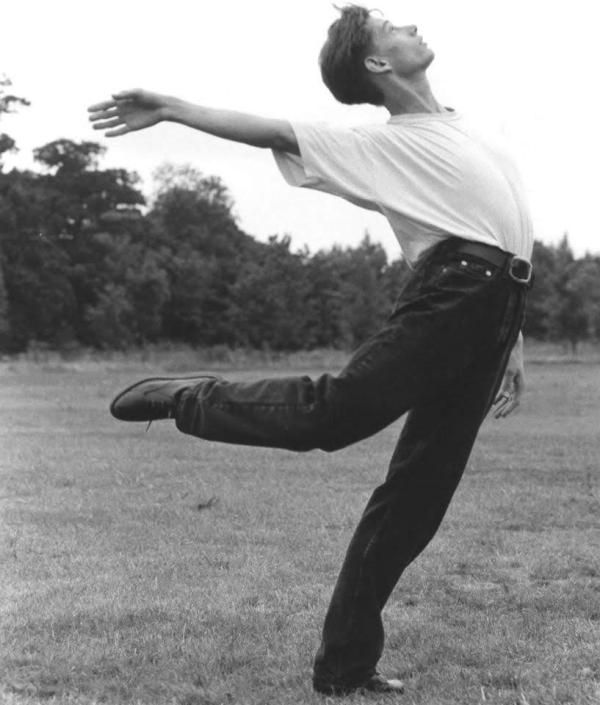 A black and white image of a young male dancer aged about 24 in a field grass under foot and trees in the background. The dancer is dressed in a white t-shirt and black jeans. His body is arched like a bow and his front right leg is bent behind him with his foot at knee height trailing behind and his front right arm reaching backwards just above shoulder height as he looks upwards at the sky.