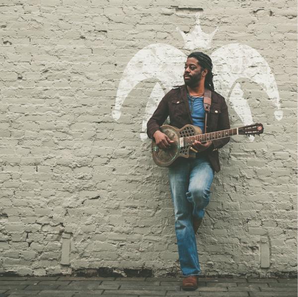 Someone posing against a whitewashed wall holing a guitar