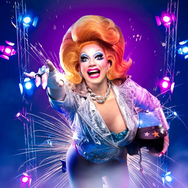 Ginger Johnson in a signature tall ginger wig wearing a space aged silver jacket, holding a helmet under her left arm, reaching for the camera.