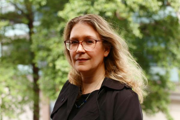 Author Sarah Brookes looks into the camera in front of a scenic woodland backdrop.