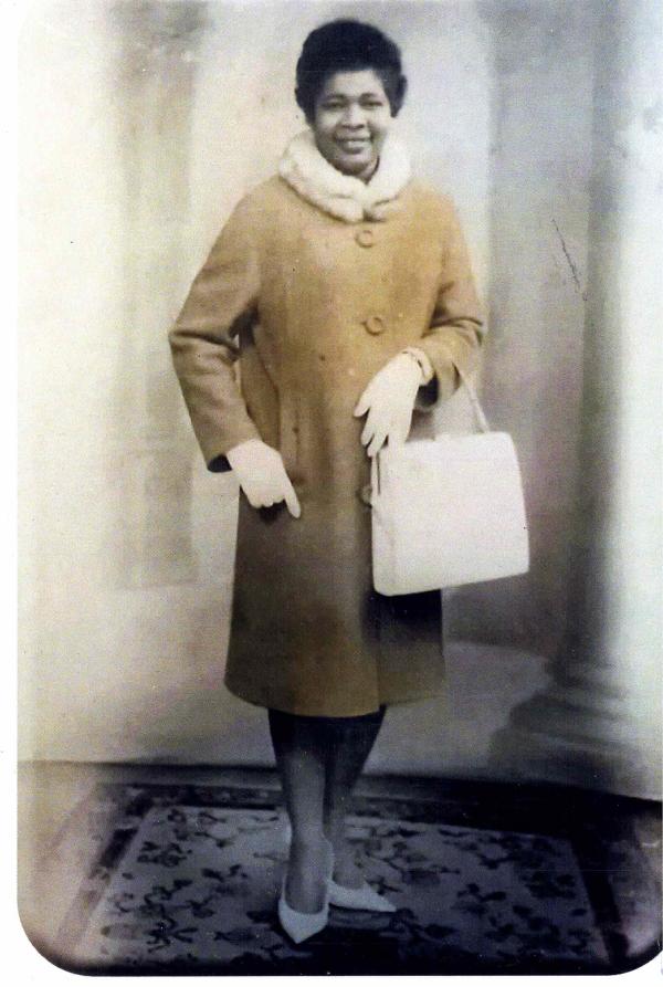 A lady in a yellow coat and white gloves and shoes smiling at the camera holding a handbag
