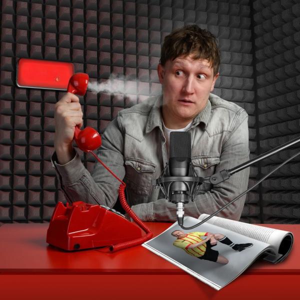 Man in recording booth in front of a recording microphone holding a wired telephone with steam coming out of it away from his head. A magazine open on a page of a kneeling footballer is open next to him.