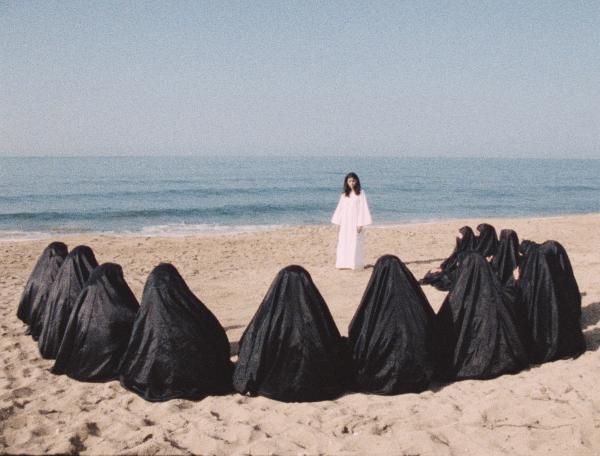 A Woman in white stands on a beach within a semicircle of 12 women in niqabs