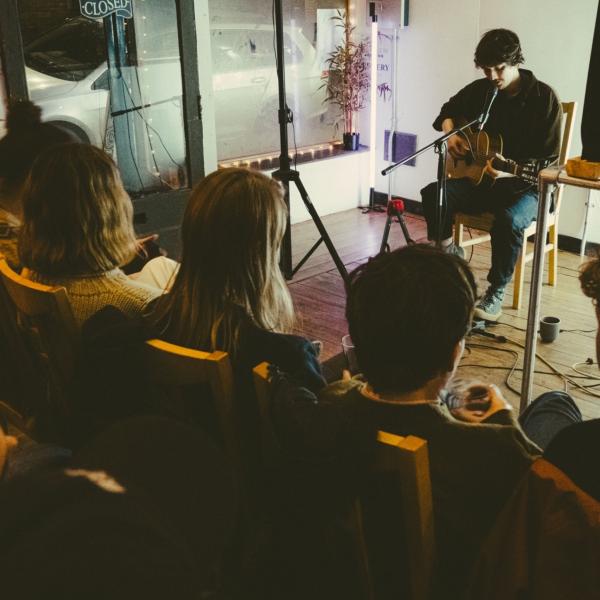 Musician Chris Brain sits in a small and intimate performance space, playing an acoustic guitar. The photo is taken from just behind a small audience that sits just in front of Chris, listening intently.