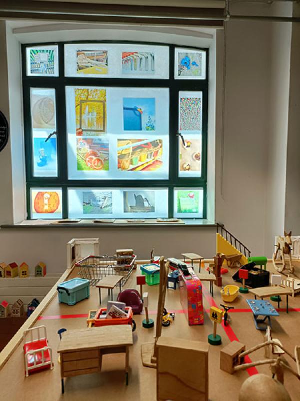 The photograph shows a table with miniature everyday objects displayed. In the background is a window with transparent photographs displayed giving a stained glass window effect.