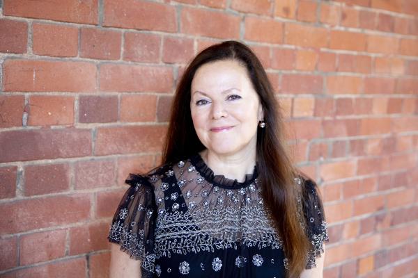 A white woman in her mid 60s. She has long straight dark hair, she is smiling, and is wearing a black sheer top with white details. There is a red brick wall behind her.
