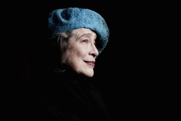 A white woman in her late 70s with grey hair. She is looking to the right and smiling, wearing a blue beret style hat and red lipstick.