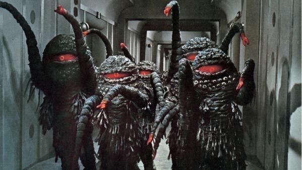 A screenshot from the film The Green Slime showing several aliens inside a space station. The aliens have one giant red eye and long tentacles